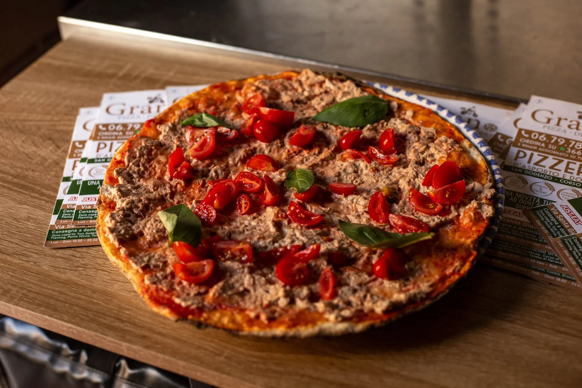 Pizza romana con pomodorini e basilico sul banco di Grano, accanto ai volantini della pizzeria